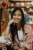 A young woman with long dark hair is smiling and resting her head on her hand. She is wearing a white top and a pink jacket. In front of her is a white bag. There is a red microphone on a stand to the left, and shelves with various items in the background.