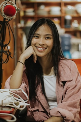 A young woman with long dark hair is smiling and resting her head on her hand. She is wearing a white top and a pink jacket. In front of her is a white bag. There is a red microphone on a stand to the left, and shelves with various items in the background.
