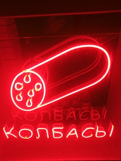 A glowing red neon sign depicting a sausage with several circular cuts shown in detail. Below the sausage image, there is text written in Cyrillic script, which is also illuminated in bright red neon.