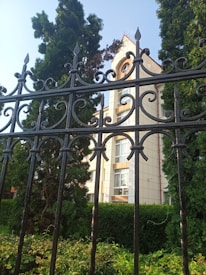 Ornate black wrought iron fence in the foreground with intricate scrollwork pattern. Behind the fence, a building with light-colored walls and multiple windows is partially visible, surrounded by tall evergreen trees and lush greenery under a clear blue sky.