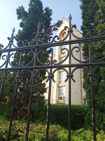 Ornate black wrought iron fence in the foreground with intricate scrollwork pattern. Behind the fence, a building with light-colored walls and multiple windows is partially visible, surrounded by tall evergreen trees and lush greenery under a clear blue sky.