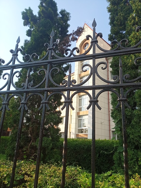 Ornate black wrought iron fence in the foreground with intricate scrollwork pattern. Behind the fence, a building with light-colored walls and multiple windows is partially visible, surrounded by tall evergreen trees and lush greenery under a clear blue sky.