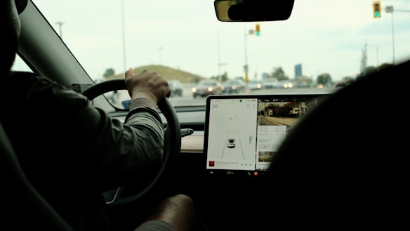 View from inside a car showing a driver with hands on the steering wheel, focusing on the dashboard and an integrated touchscreen displaying navigation information. The car appears to be stopped at an intersection, with visible traffic lights ahead.