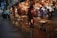 A cozy outdoor café adorned with vibrant flowers and string lights. Wooden chairs and tables are arranged neatly under a pergola, creating a warm and inviting ambiance. The atmosphere suggests a quiet, intimate dining experience on a city street during the evening.