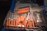 A metal tray filled with multiple syringes containing an orange substance. The syringes are of various sizes and are predominantly arranged with the needle-end facing downward. Adjacent to the syringes, there are plastic bags and medical instruments, suggesting a medical or laboratory setting.