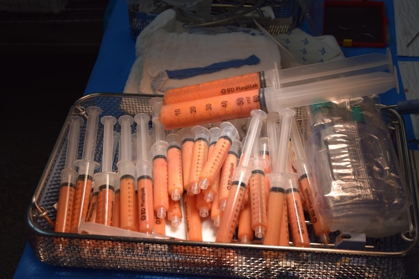 A metal tray filled with multiple syringes containing an orange substance. The syringes are of various sizes and are predominantly arranged with the needle-end facing downward. Adjacent to the syringes, there are plastic bags and medical instruments, suggesting a medical or laboratory setting.