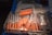 A metal tray filled with multiple syringes containing an orange substance. The syringes are of various sizes and are predominantly arranged with the needle-end facing downward. Adjacent to the syringes, there are plastic bags and medical instruments, suggesting a medical or laboratory setting.