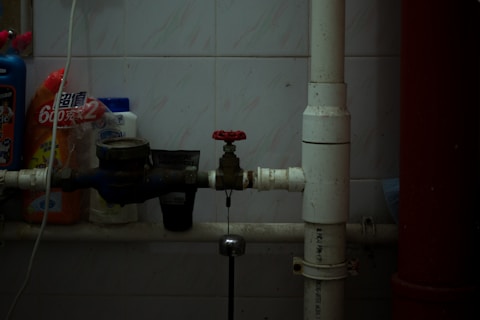 A dimly lit utility area with pipes, a valve, and various cleaning products lined up against a tiled wall. The central focus is on a red-handled valve attached to white and blue pipes running horizontally. Several bottles of cleaning supplies with Asian script are visible, suggesting a storage or maintenance corner.