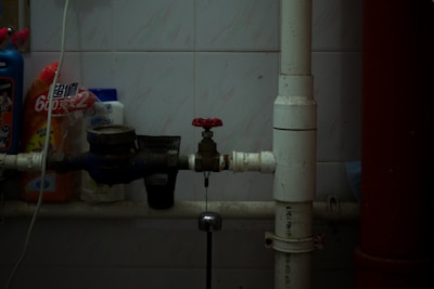 A dimly lit utility area with pipes, a valve, and various cleaning products lined up against a tiled wall. The central focus is on a red-handled valve attached to white and blue pipes running horizontally. Several bottles of cleaning supplies with Asian script are visible, suggesting a storage or maintenance corner.