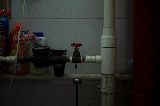 A dimly lit utility area with pipes, a valve, and various cleaning products lined up against a tiled wall. The central focus is on a red-handled valve attached to white and blue pipes running horizontally. Several bottles of cleaning supplies with Asian script are visible, suggesting a storage or maintenance corner.