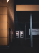A modern architectural scene featuring a building with large yellow panels. Two glass doors are visible, reflecting a street scene. A gray pillar stands in the foreground along with a sign that has text in a foreign language.