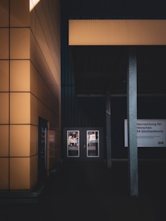 A modern architectural scene featuring a building with large yellow panels. Two glass doors are visible, reflecting a street scene. A gray pillar stands in the foreground along with a sign that has text in a foreign language.