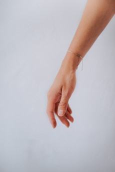 A close-up of a human hand gracefully extended downward against a light, neutral background. The hand is wearing a delicate, thin bracelet.