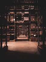 A long warehouse aisle is lined with tall metal shelves filled with large, stacked boxes and pallets. The space is dimly lit, creating a tunnel effect down the aisle with visible symmetry and perspective extending into the distance.