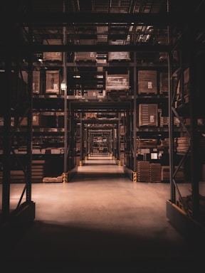 A long warehouse aisle is lined with tall metal shelves filled with large, stacked boxes and pallets. The space is dimly lit, creating a tunnel effect down the aisle with visible symmetry and perspective extending into the distance.
