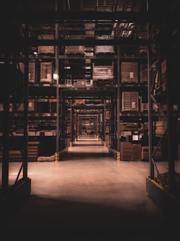 A long warehouse aisle is lined with tall metal shelves filled with large, stacked boxes and pallets. The space is dimly lit, creating a tunnel effect down the aisle with visible symmetry and perspective extending into the distance.