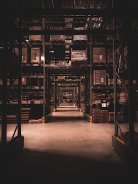 A long warehouse aisle is lined with tall metal shelves filled with large, stacked boxes and pallets. The space is dimly lit, creating a tunnel effect down the aisle with visible symmetry and perspective extending into the distance.