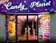 A brightly lit candy store entrance with a neon purple sign reading 'Candy Planet'. The interior is filled with various colorful sweets and candies displayed on shelves. There are vibrant lights and candy-related decorations scattered throughout.