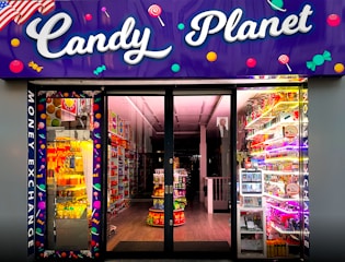 A brightly lit candy store entrance with a neon purple sign reading 'Candy Planet'. The interior is filled with various colorful sweets and candies displayed on shelves. There are vibrant lights and candy-related decorations scattered throughout.