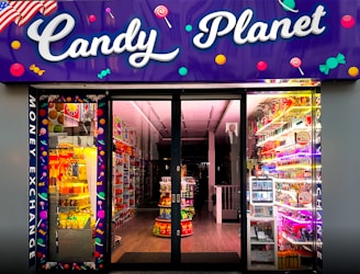 A brightly lit candy store entrance with a neon purple sign reading 'Candy Planet'. The interior is filled with various colorful sweets and candies displayed on shelves. There are vibrant lights and candy-related decorations scattered throughout.