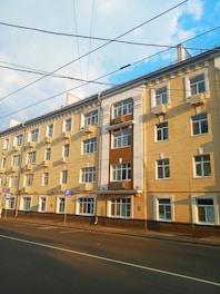 A multi-story residential building with a series of windows and balconies. The facade of the building is painted in shades of beige and brown, with white decorative elements. Several air conditioning units are mounted near the windows. The street in front of the building is empty, and overhead wires are visible against a partly cloudy sky.