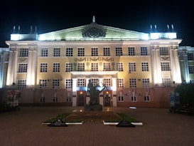 A large, illuminated classical-style building with symmetrical architecture, featuring multiple rows of windows and prominent rectangular columns. The building, possibly an institutional or governmental structure, is lit up at night, highlighting the intricate details of its facade. In front of the entrance, there is a statue or monument, which stands on a pedestal on a tiled plaza.