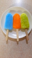 Three colorful popsicles are placed on a white plate. They are arranged in a row with the first being blue, the second orange, and the third green. Each popsicle has a wooden stick and they are set against a light-colored, slightly textured surface.