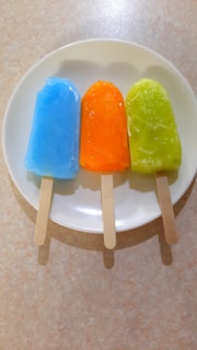 Three colorful popsicles are placed on a white plate. They are arranged in a row with the first being blue, the second orange, and the third green. Each popsicle has a wooden stick and they are set against a light-colored, slightly textured surface.