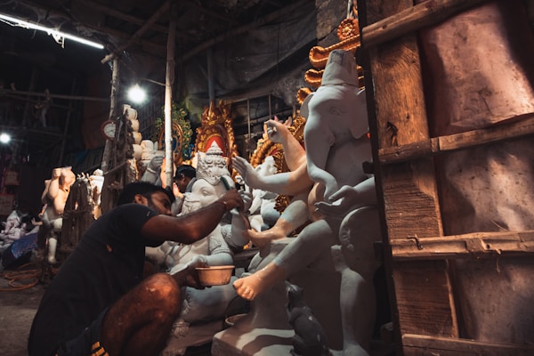 An artisan is meticulously painting a large statue of the Hindu deity Ganesha in a workshop. The setting is vibrant with multiple unfinished idols visible in the background. The workshop has a rustic feel, with dim lighting and wooden scaffolding.