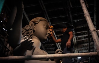 A craftsman meticulously works on a large clay sculpture of a deity inside a dimly lit workshop. The sculpture has intricate details, including flowing hair and a serene facial expression. Wooden scaffolding surrounds the scene, and the craftsman uses a cloth to smooth the surface.