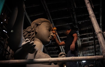 A craftsman meticulously works on a large clay sculpture of a deity inside a dimly lit workshop. The sculpture has intricate details, including flowing hair and a serene facial expression. Wooden scaffolding surrounds the scene, and the craftsman uses a cloth to smooth the surface.