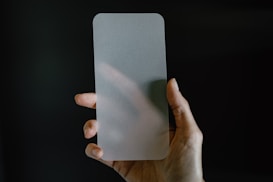 A hand is holding a frosted glass screen protector against a dark background. The glass has rounded corners and appears translucent, partially obscuring the hand behind it.