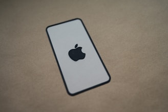 A minimalist image of a paper cutout shaped like a smartphone, featuring a prominent apple logo in the center, placed on a plain brown background.