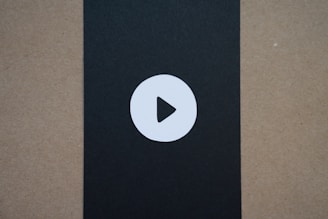 A simple circular play button icon is centered on a black vertical strip that contrasts with the surrounding brownish-beige background.