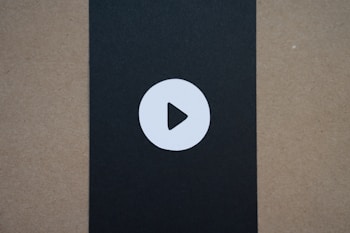 A simple circular play button icon is centered on a black vertical strip that contrasts with the surrounding brownish-beige background.