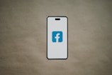 A smartphone is depicted with a white screen displaying a blue logo with the letter 'f', set against a plain brown background.