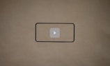 A blank brown background with a black rectangle outline in the center. Inside the outline, there is a light gray play button icon.