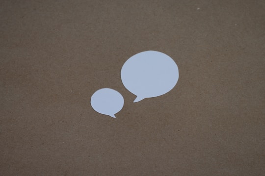 Two white paper cutouts shaped like speech bubbles are placed on a brown, textured background. The larger bubble is on the right and the smaller bubble is towards the left.