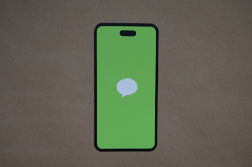 A smartphone design with a green screen and a white speech bubble icon in the center is placed on a plain brown surface. The top of the phone features a small notch.