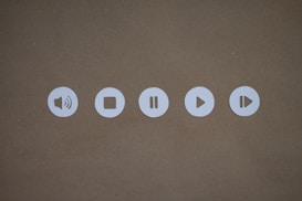 Five circular white icons on a brown background, each representing sound and playback controls. These include a speaker, stop, pause, play, and fast-forward symbol.