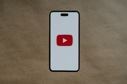 A smartphone-like rectangular shape is centered on a brown textured background. The shape has a white screen with rounded edges and features a prominent red play button inside a white triangle in the center, resembling the YouTube logo.