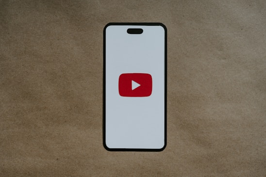 A smartphone-like rectangular shape is centered on a brown textured background. The shape has a white screen with rounded edges and features a prominent red play button inside a white triangle in the center, resembling the YouTube logo.