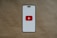 A smartphone-like rectangular shape is centered on a brown textured background. The shape has a white screen with rounded edges and features a prominent red play button inside a white triangle in the center, resembling the YouTube logo.