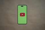 A smartphone silhouette with a bright green screen displays a red play button symbol in the center, resembling the YouTube logo. The background is a plain, neutral brown surface.