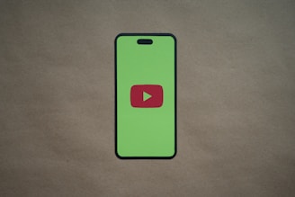 A smartphone silhouette with a bright green screen displays a red play button symbol in the center, resembling the YouTube logo. The background is a plain, neutral brown surface.
