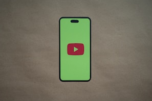 A smartphone silhouette with a bright green screen displays a red play button symbol in the center, resembling the YouTube logo. The background is a plain, neutral brown surface.