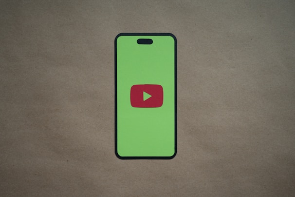 A smartphone silhouette with a bright green screen displays a red play button symbol in the center, resembling the YouTube logo. The background is a plain, neutral brown surface.