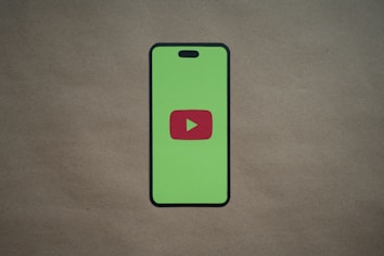 A smartphone silhouette with a bright green screen displays a red play button symbol in the center, resembling the YouTube logo. The background is a plain, neutral brown surface.