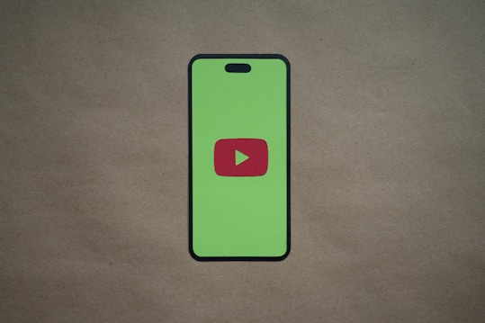 A smartphone silhouette with a bright green screen displays a red play button symbol in the center, resembling the YouTube logo. The background is a plain, neutral brown surface.