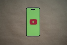 A smartphone silhouette with a bright green screen displays a red play button symbol in the center, resembling the YouTube logo. The background is a plain, neutral brown surface.